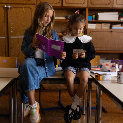 Deux fillettes assises sur une table dans une salle de classe, l'une tient un cahier, l'autre une carte de conversation